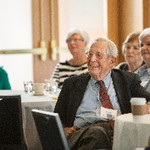 A smiling man is seated at a table, while women are seated behind him and out of focus